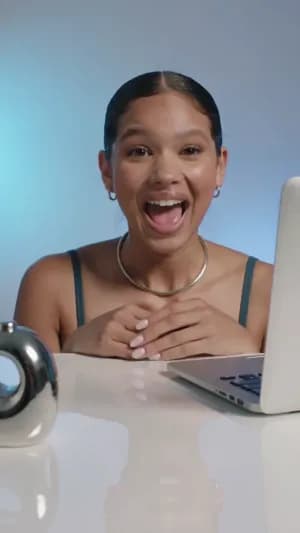 A vertical talking head UGC video of a young creative woman sitting at a clean desk with a laptop, reacting excitedly while talking to the camera. Modern Y3K Pinterest aesthetic, chrome desk accessories, soft blue lighting, glossy makeup, sleek hair, silver jewelry, authentic TikTok creator vibe, natural hand gestures, slight camera shake. She clearly says: "YOUR TEXT HERE"